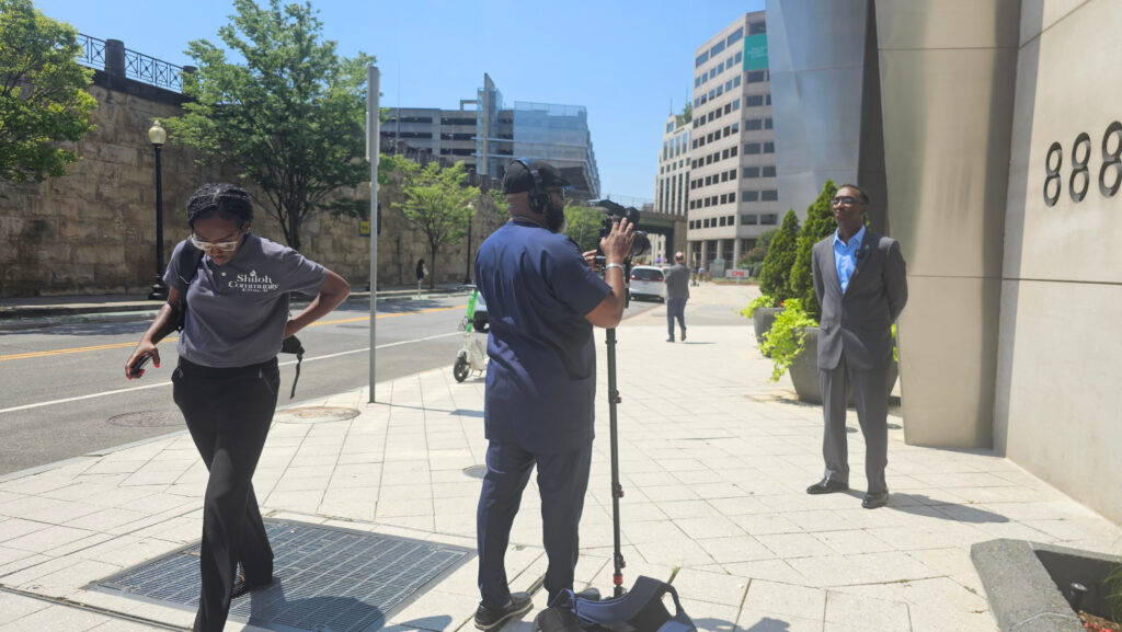 Documentarian, and Founder/Director of the Peoples Justice Council, Michael Malcom sets up a tripod to interview Pastor Timothy Williams of Shiloh Community Center.  Pastor Williams, daughter and Shiloh Community Center Executive Assistant, Melissa Williams walk to the left of Michael Malcom. 